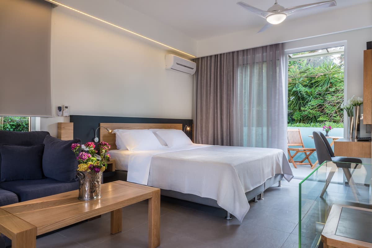 A modern hotel room with a bed, a sofa, a dining table, and a balcony with a view of greenery.