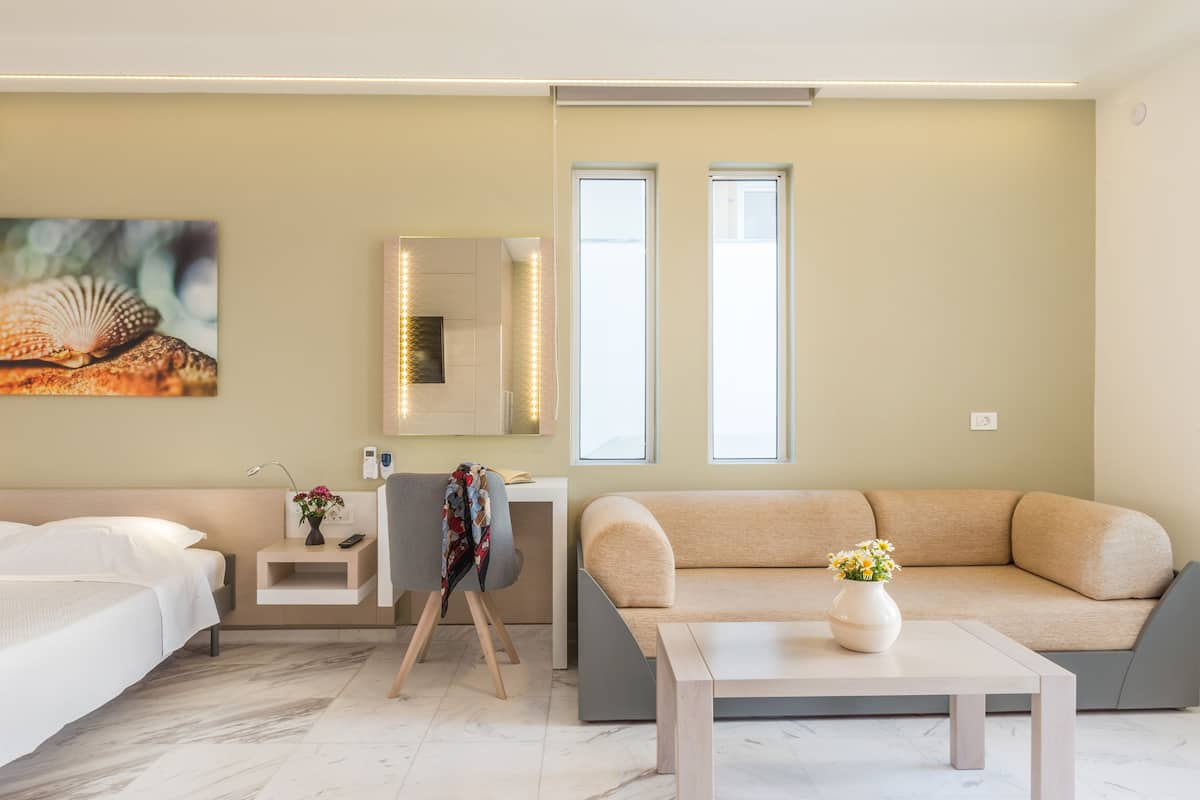 A modern hotel room with a bed, a sofa, a small table, a nightstand, a mirror, and a painting on the wall.