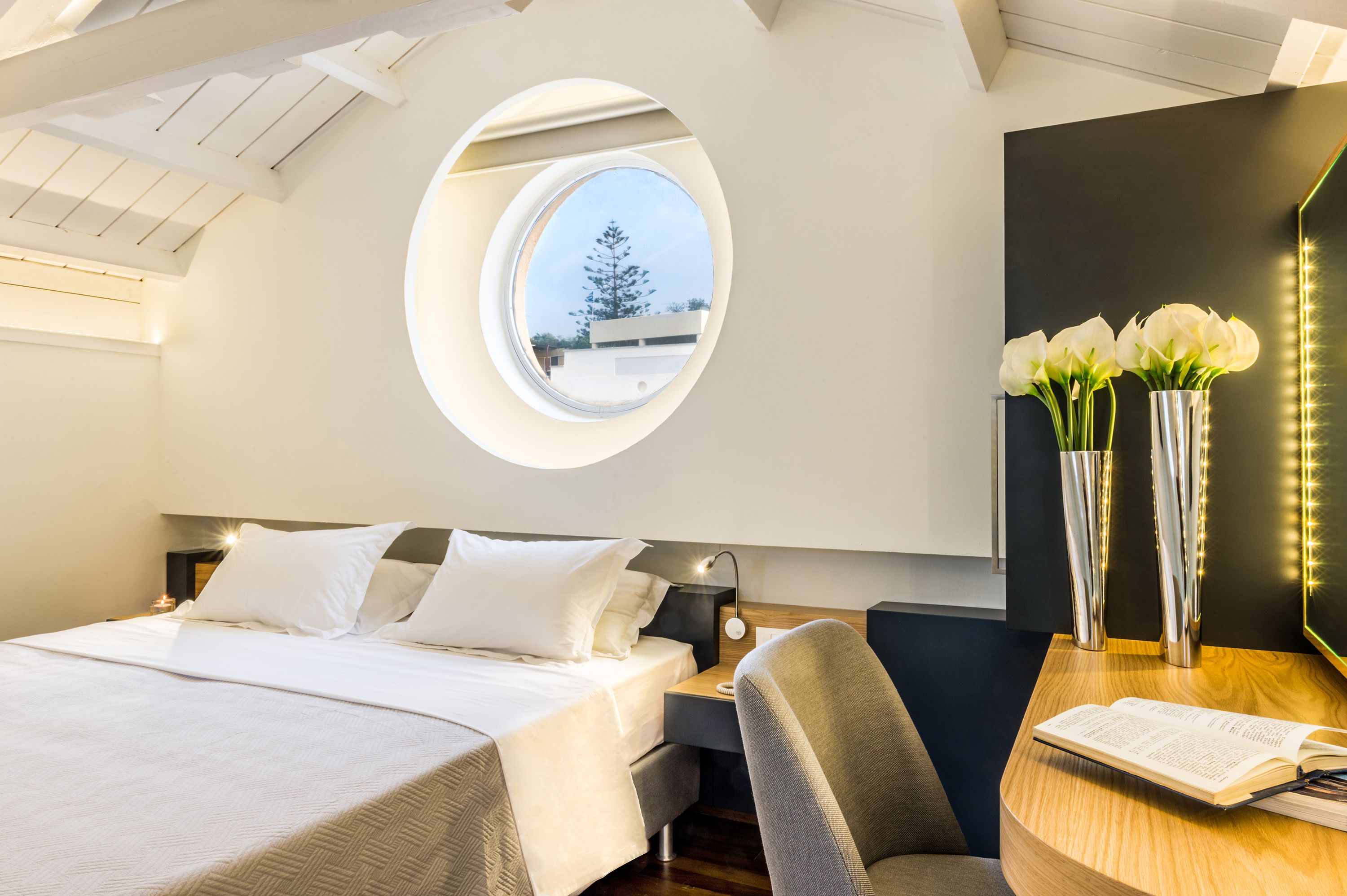 Habitació d'hotel moderna amb un fanal, una finestra rodona, un llit amb roba de llit blanca, una taula de treball amb una làmpada i un vas amb flors blanques.