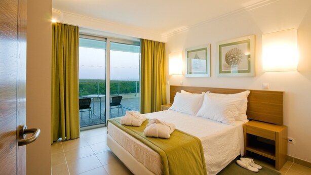 A hotel room with a bed, a nightstand, a lamp, and a view of the outdoors. - Monte Gordo Hotel Apartamentos & Spa (Vila Real Santo Antonio)