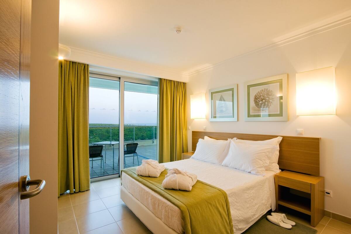 Ein Hotelzimmer mit Bett, Nachttisch, Lampe und Blick ins Freie.