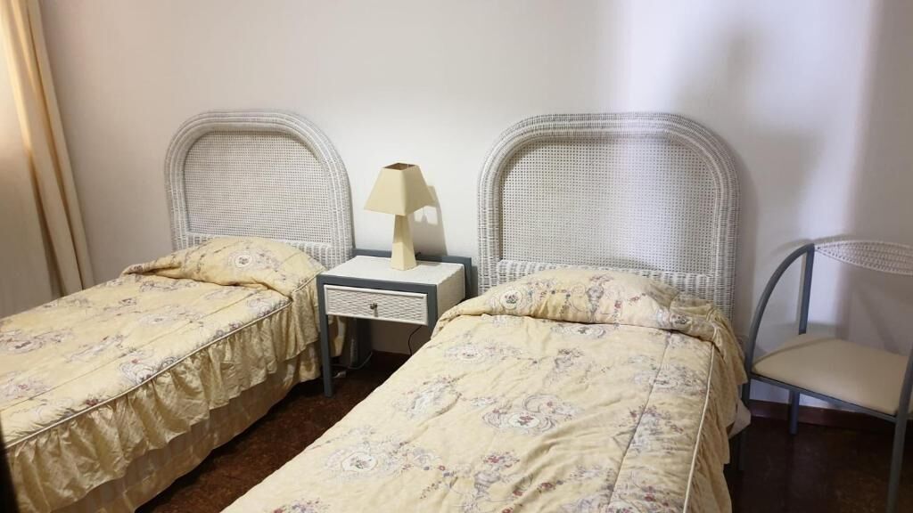 Una habitación con dos camas individuales, una mesita de noche con lámpara y una silla.