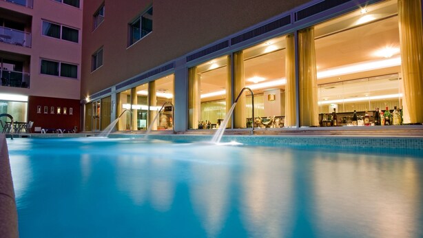 Una piscina interior con una fuente de agua, contigua a un edificio con ventanas amplias y un interior bien iluminado.