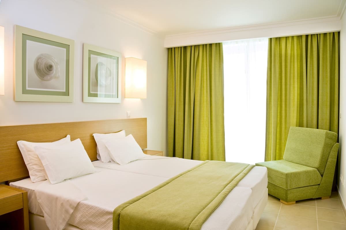 Ein Hotelzimmer mit einem großen Bett, zwei Nachttischen, einem grünen Sessel und zwei gerahmten Kunstwerken an der Wand.