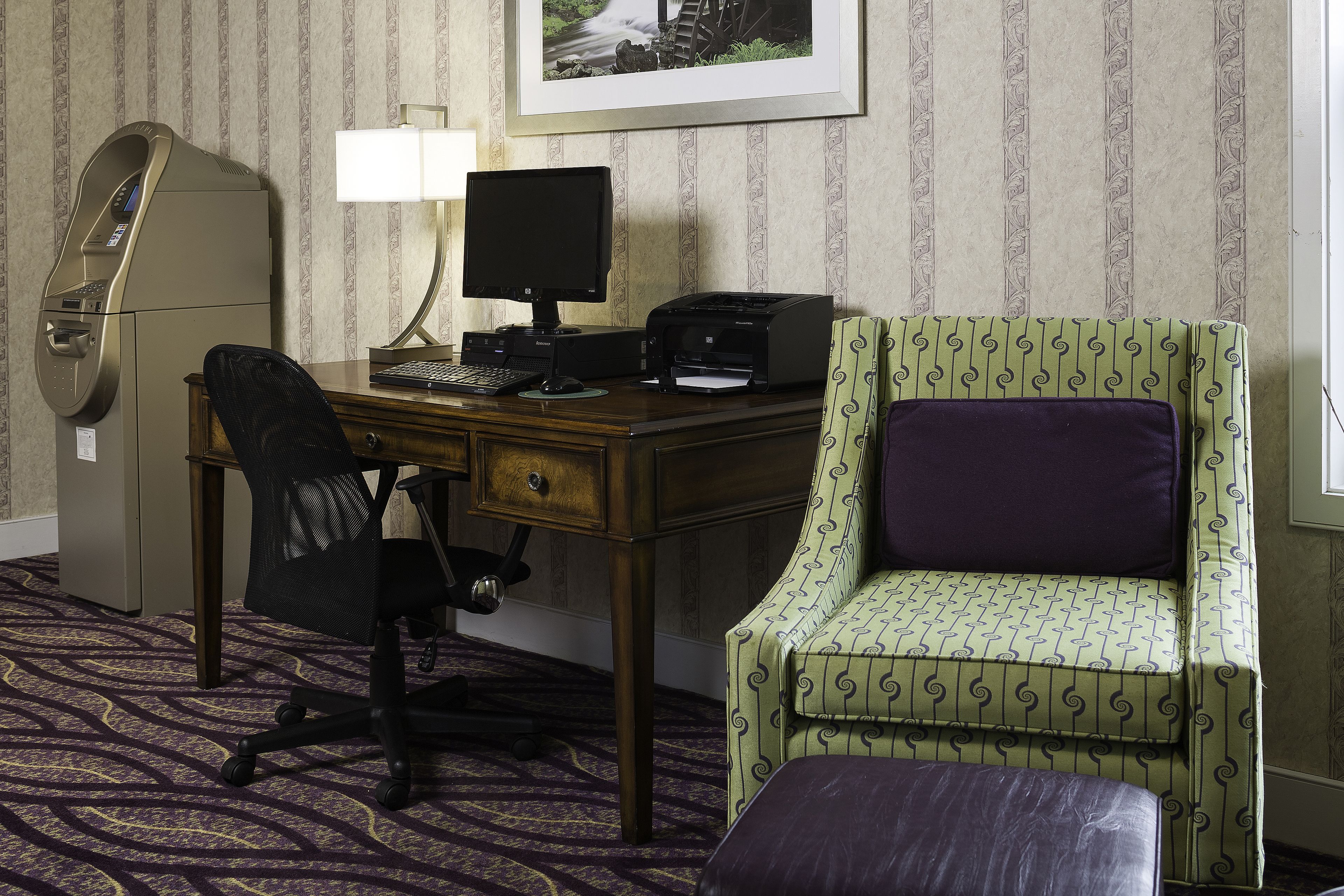 A hotel room with a desk, computer, printer, and a green patterned armchair.