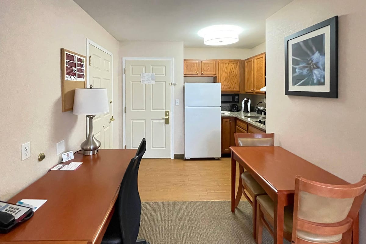 A hotel room with a dining table, chairs, a refrigerator, wooden cabinets, and a framed picture on the wall.
