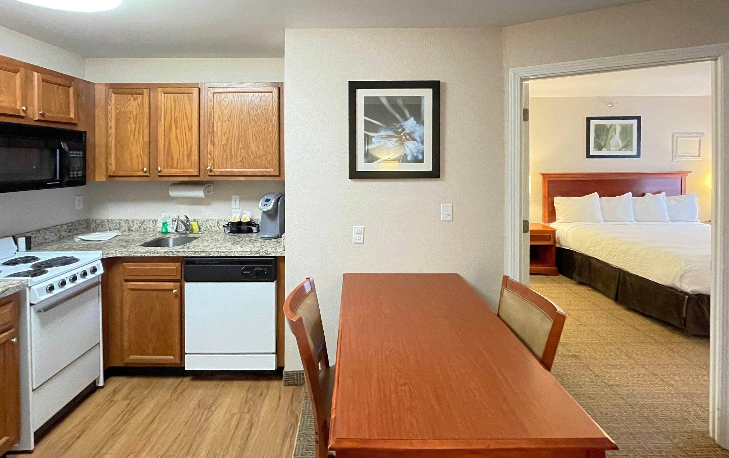 A compact kitchen with a stove, microwave, and dishwasher, a dining table with chairs, and a bedroom with a bed and a framed picture on the wall.