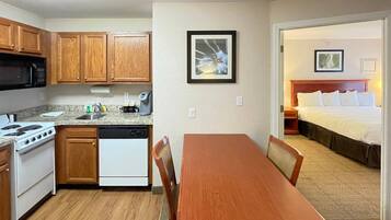 A compact kitchen with a stove, microwave, and dishwasher, a dining table with chairs, and a bedroom with a bed and a framed picture on the wall.
