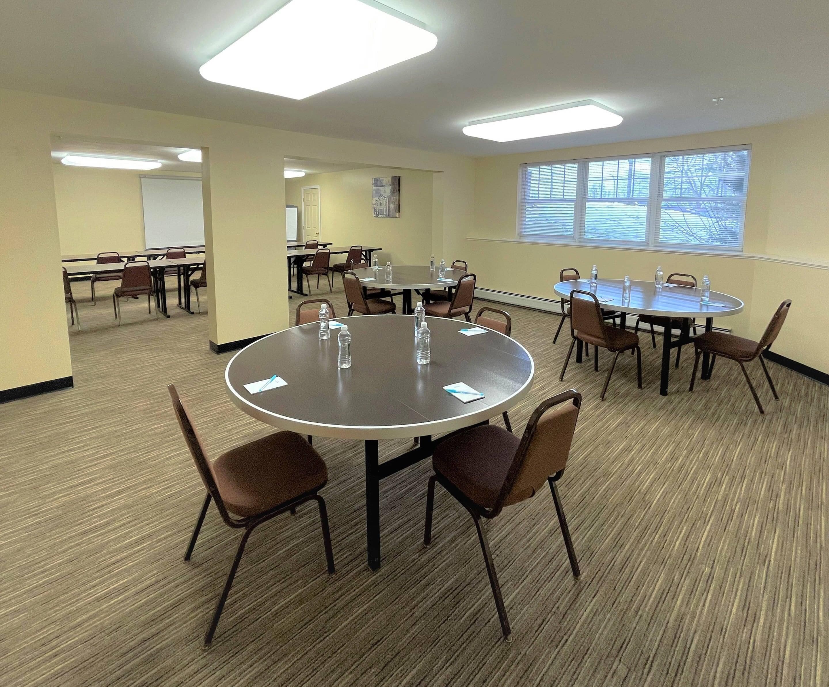 A conference room with multiple tables and chairs, carpeted floors, and large windows.