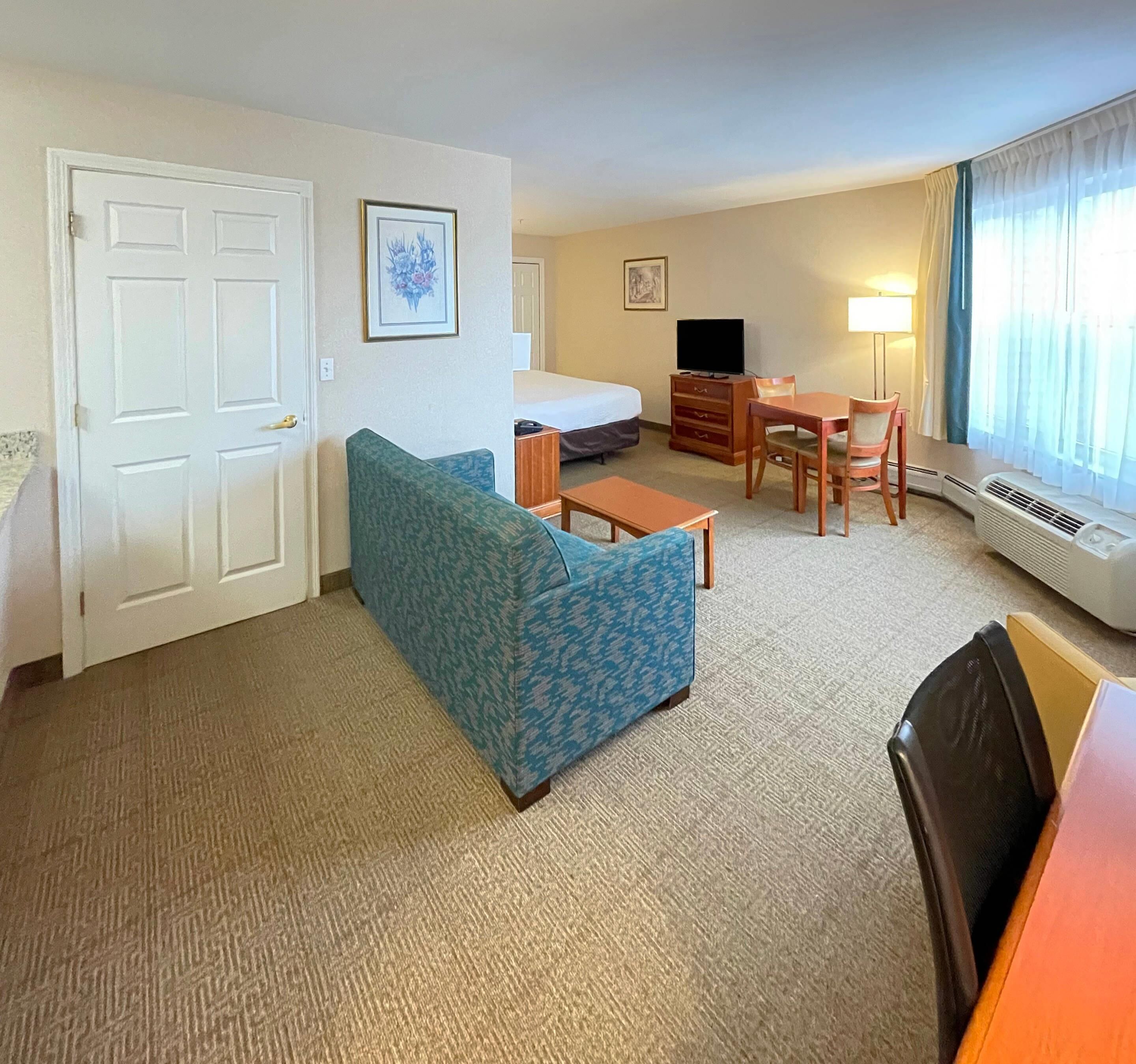Une chambre d’hôtel avec un canapé, une petite table, une télévision, un bureau et une chaise. Il y a une porte et une fenêtre avec des rideaux.