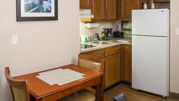 A compact kitchen with wooden cabinets, a white refrigerator, a dining table with chairs, and a framed picture on the wall.