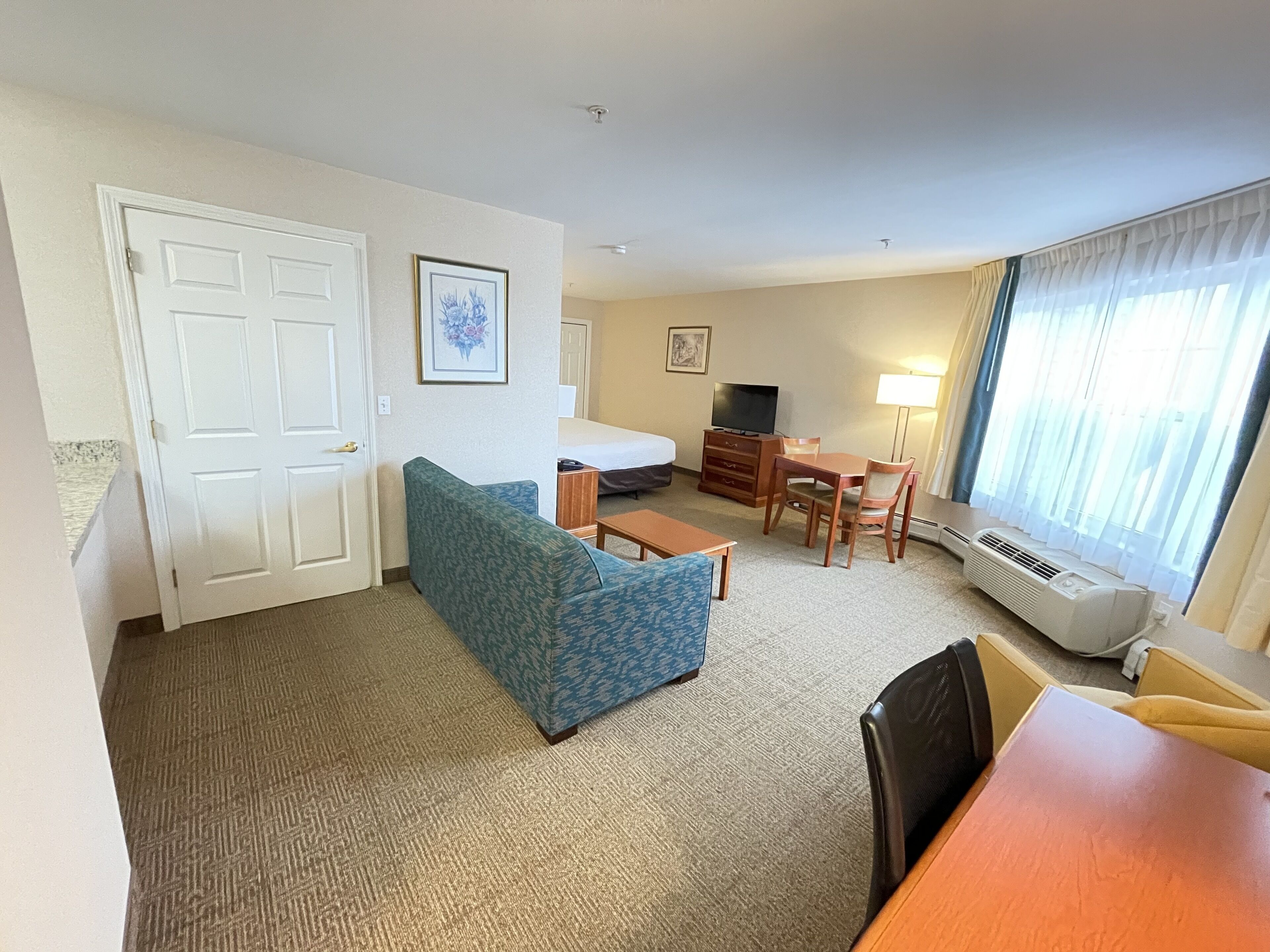 A hotel room with a blue patterned sofa, a wooden dining table with chairs, a small TV, and a bed with a white headboard.