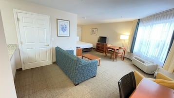 A hotel room with a blue patterned sofa, a wooden dining table with chairs, a small TV, and a bed with a white headboard.