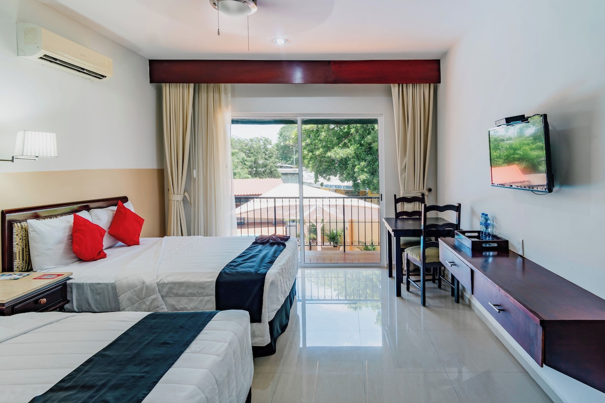 Habitación de hotel con dos camas, un escritorio, una silla, un televisor y un balcón con vistas a árboles y edificios.