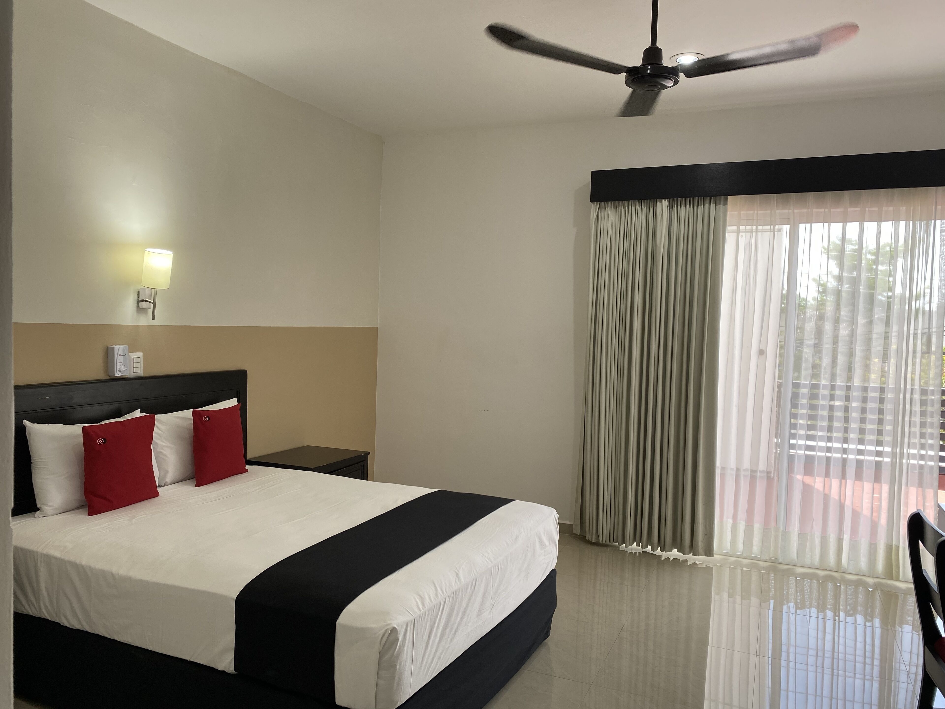 Habitación de hotel con cama, ventilador de techo, mesita de noche, lámpara y ventana con cortinas.