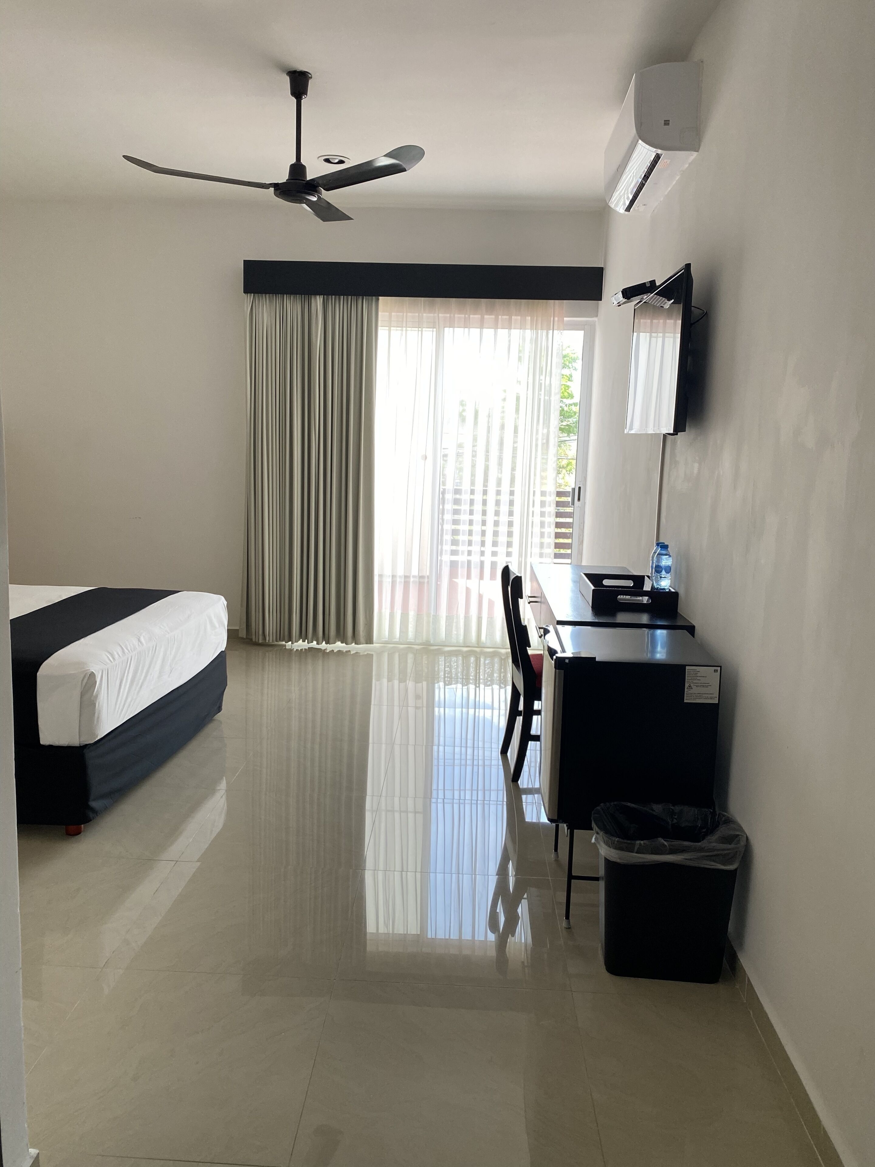 Habitación de hotel con cama, escritorio, silla, cubo de basura, ventilador de techo y unidad de aire acondicionado.