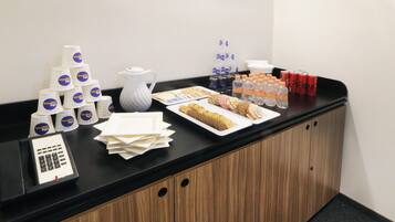 A counter with a coffee maker, stacked cups, water bottles, cookies, and a cash register.