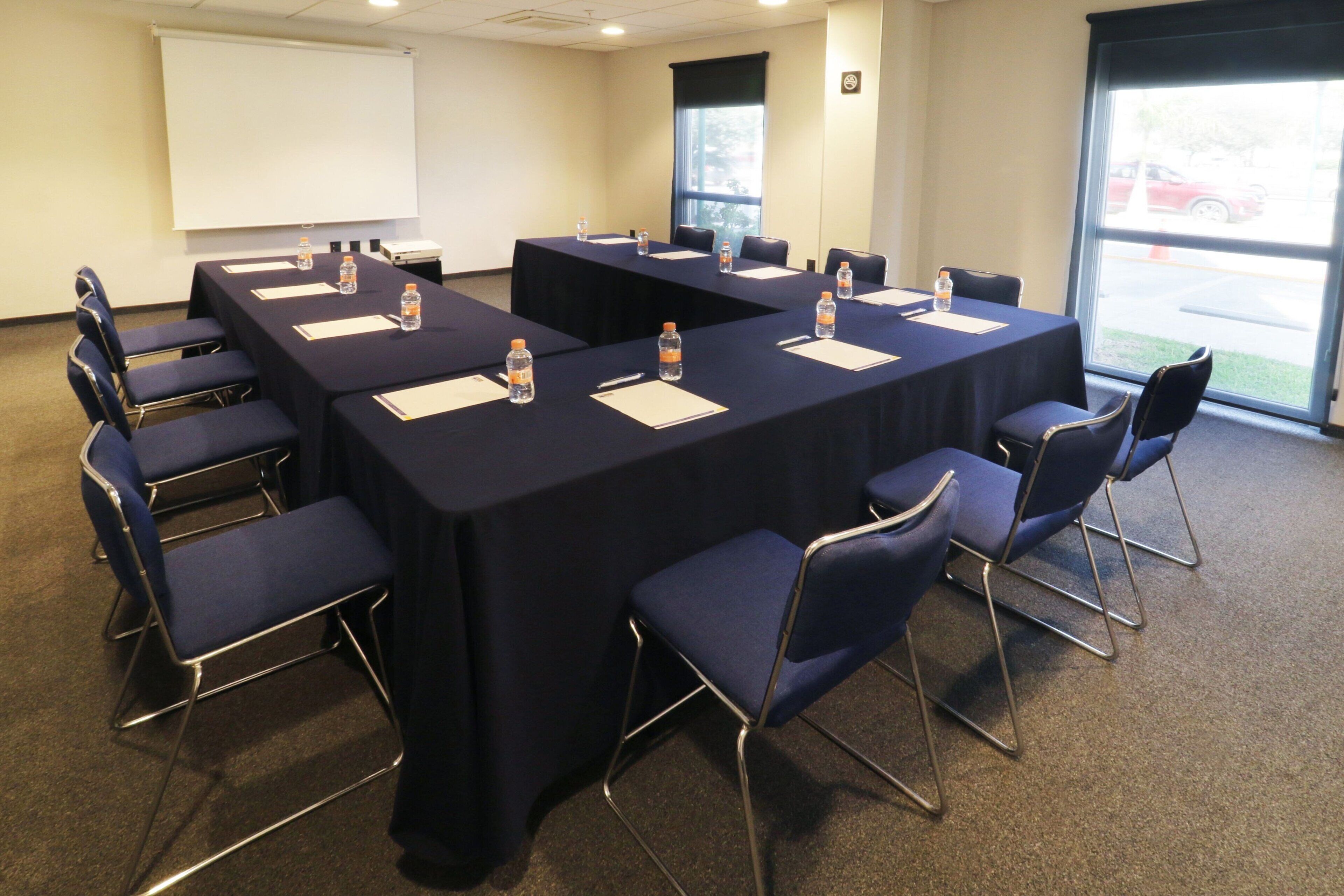 Un salón de conferencias con una gran mesa, sillas, botellas de agua y una pantalla de proyector.