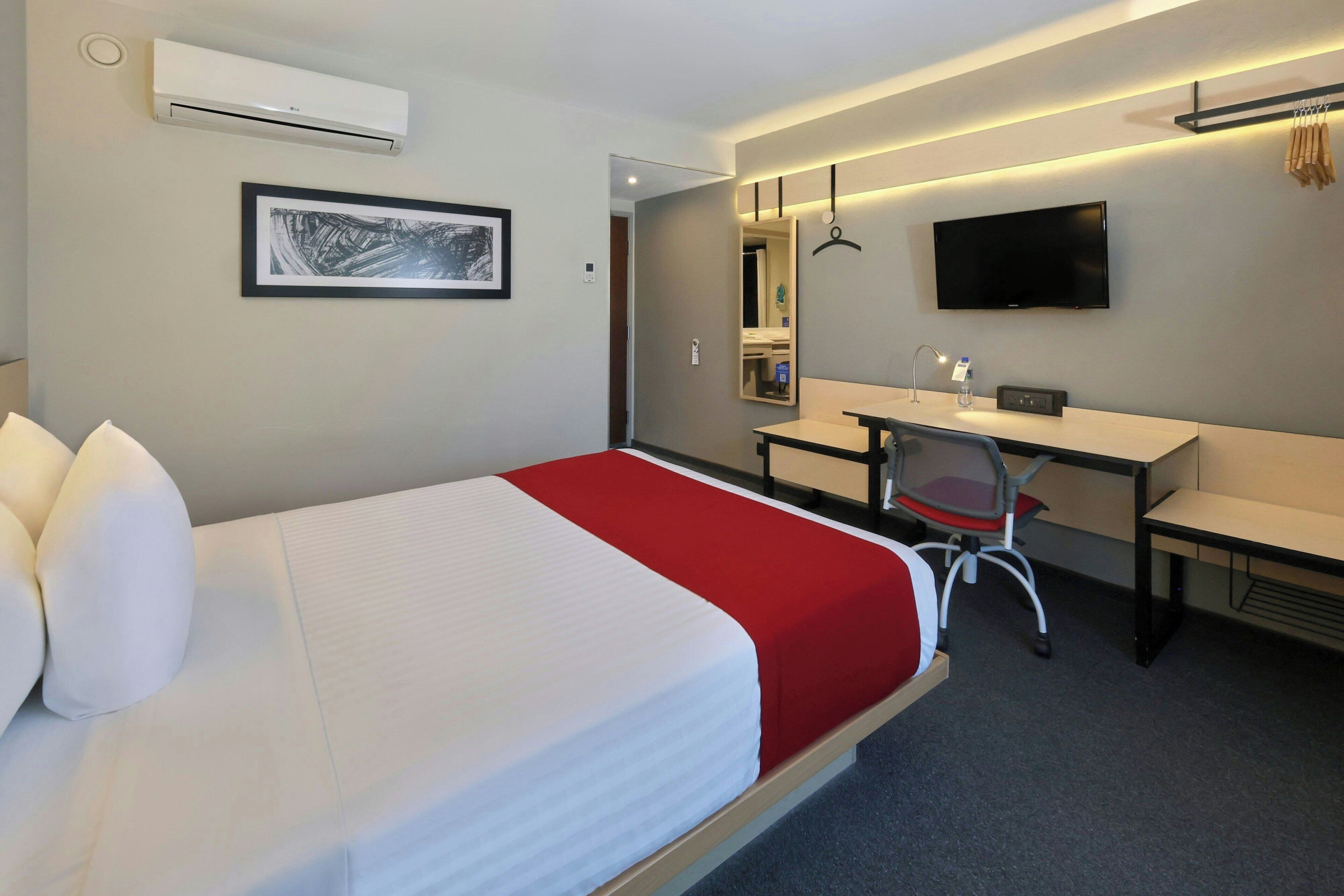 A hotel room with a bed, a desk, a chair, a TV, and an air conditioning unit.