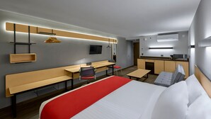 Quarto de hotel moderno com uma cama grande, uma escrivaninha, uma cadeira, um banco, uma TV e uma kitchenette.