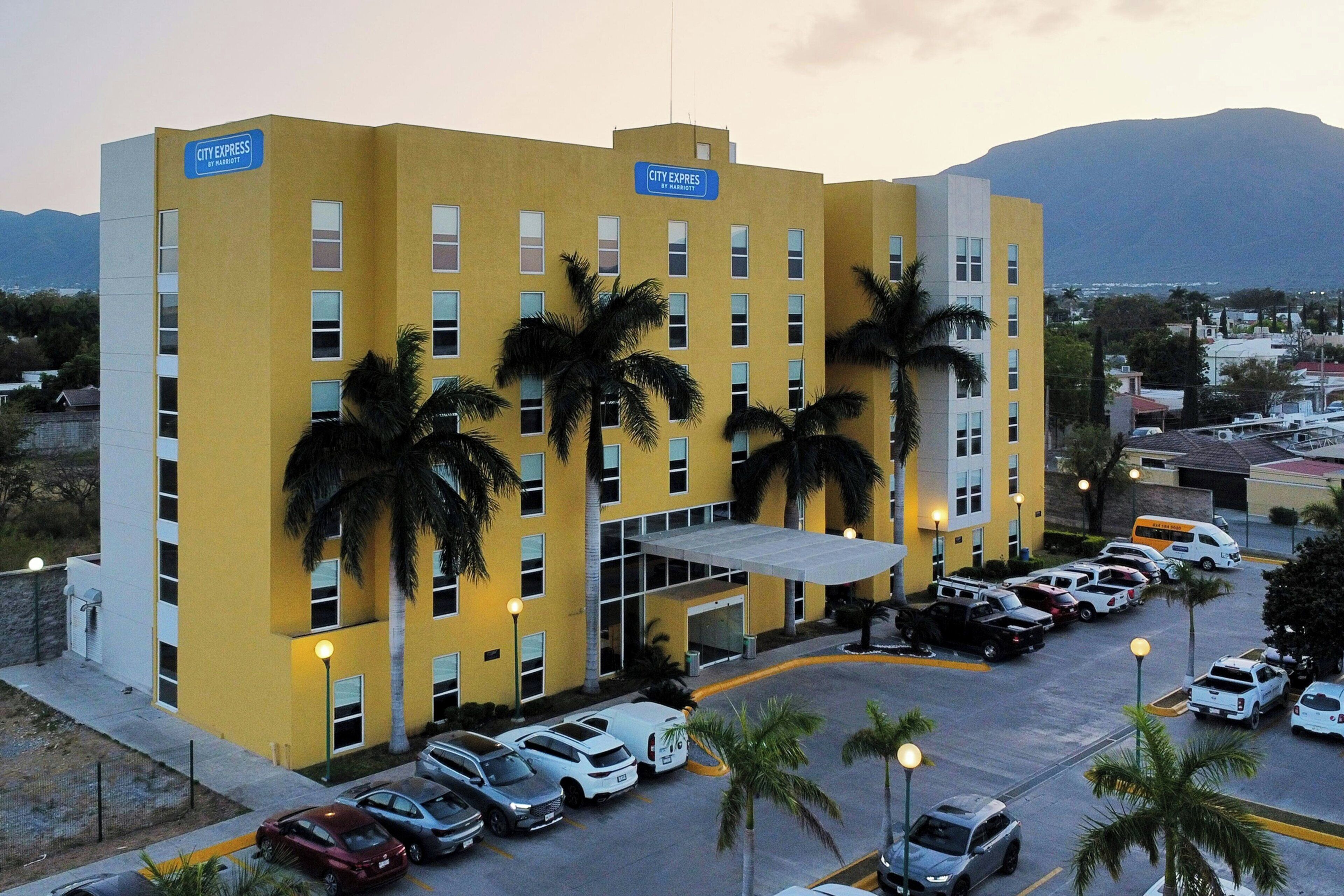 Un edificio amarillo con letrero de 'City Express', múltiples ventanas y un estacionamiento con coches.