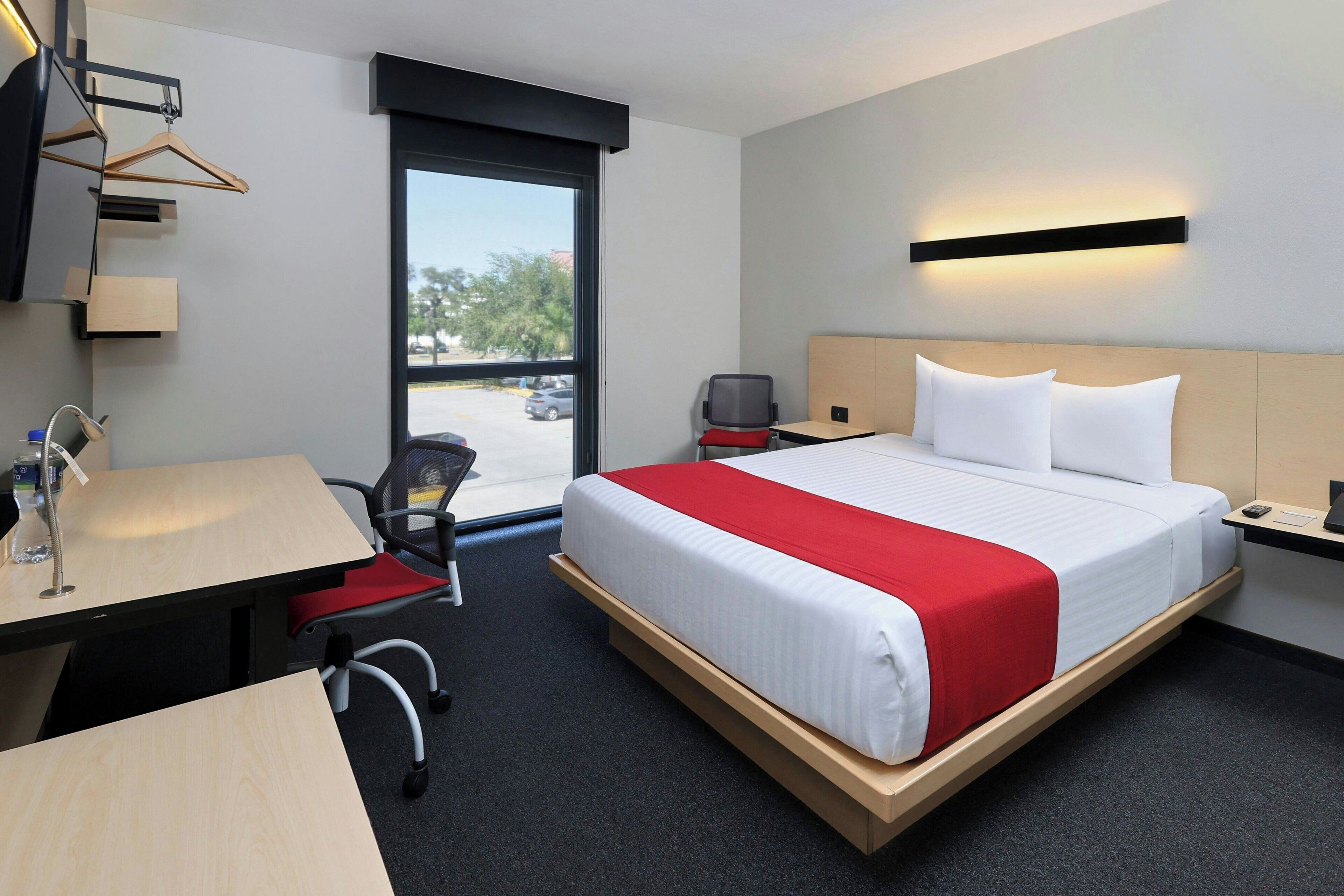 A hotel room with a bed, desk, chair, and a view of the outside.