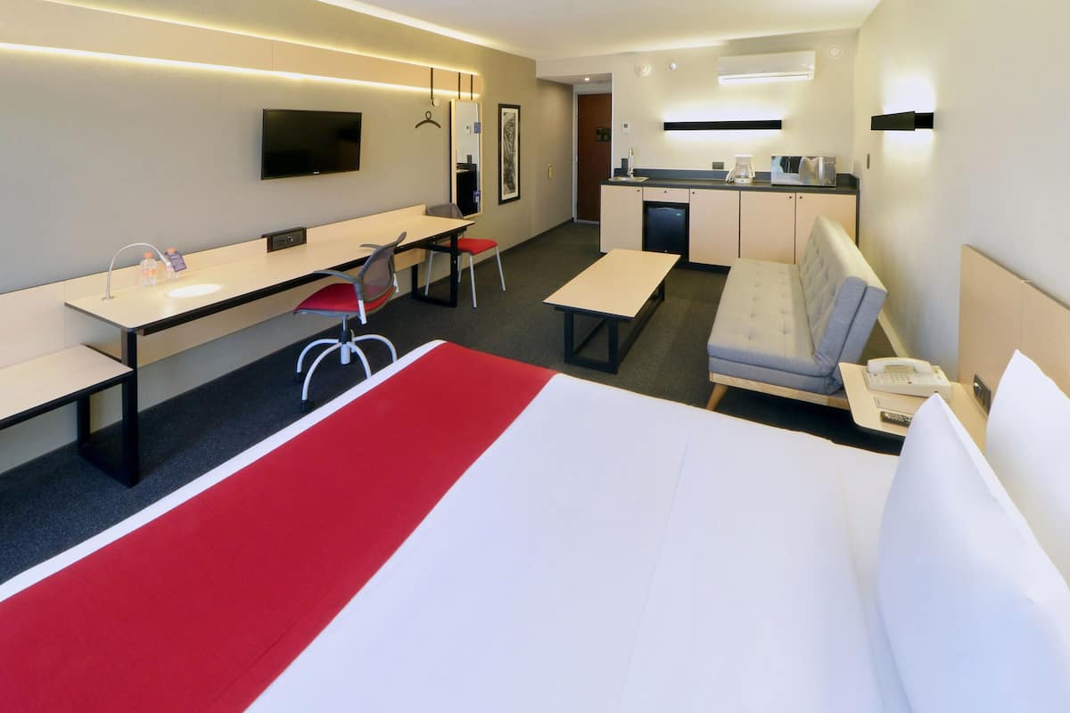A hotel room with a bed, a desk, a chair, a sofa, a small kitchenette, and a TV.