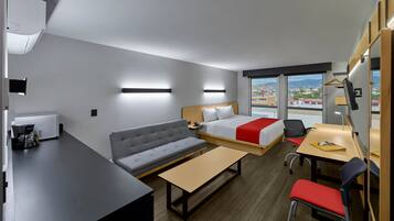 A modern hotel room with a bed, sofa, desk, and a view of the city.