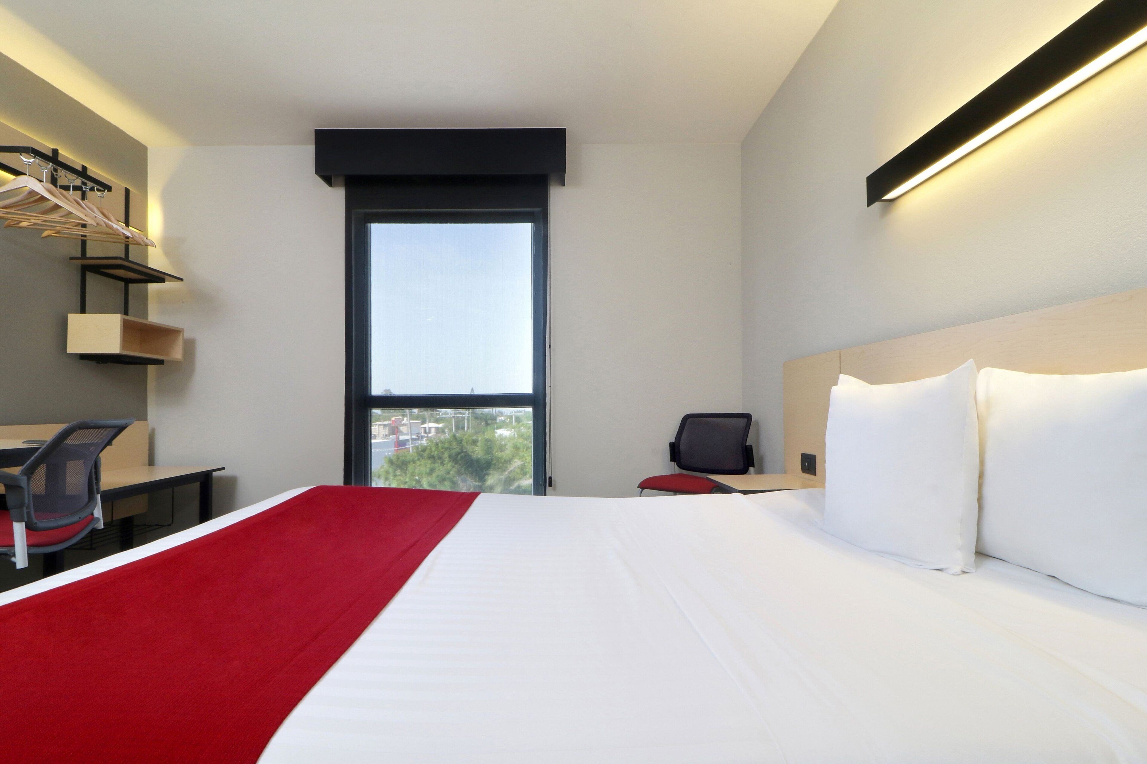 Habitación de hotel con una cama grande, un escritorio con silla, una mesita y una ventana que ofrece vistas a edificios y árboles.
