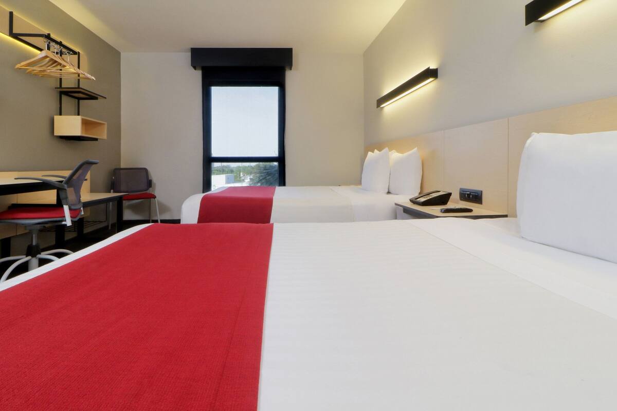 A hotel room with a bed, a desk, a chair, a window, and a nightstand with a phone.
