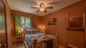 Un dormitorio con cama de madera, ventilador de techo, lámpara de noche, ventana con cortinas y una banca de madera.