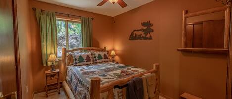 A bedroom with a wooden bed, a ceiling fan, a bedside lamp, a window with curtains, and a wooden bench.