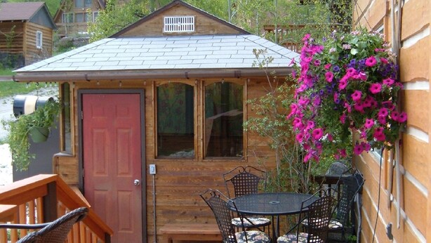 Una terraza de madera con una mesa, sillas, una puerta roja y una cesta colgante de flores.