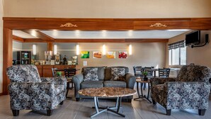 A cozy living area with patterned armchairs, a round coffee table, and a sofa. A kitchen area is visible in the background with a refrigerator and a dining table.
