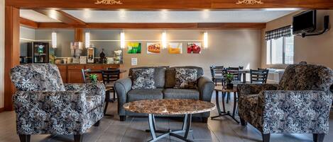 Un salon chaleureux avec des fauteuils à motifs, une table basse ronde et un canapé. On aperçoit à l’arrière-plan un coin cuisine avec un réfrigérateur et une table à manger.