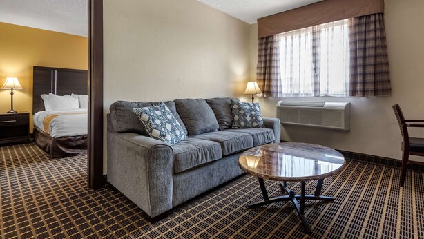 A hotel room with a bed, a gray sofa with patterned cushions, a round coffee table, and a window with plaid curtains.