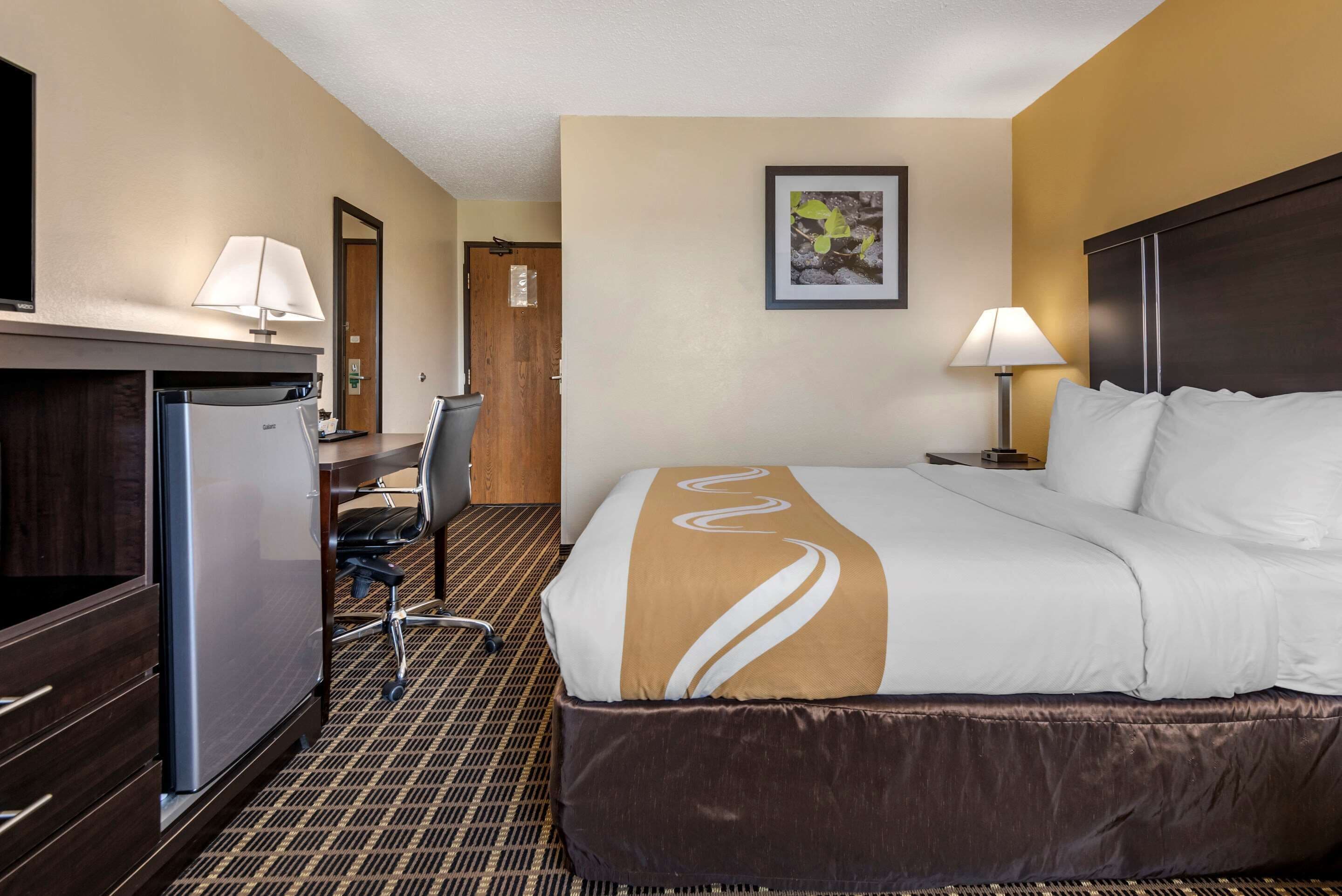 A hotel room with a bed, desk, refrigerator, and a chair.