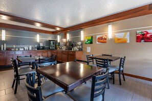 A spacious dining area with wooden tables and chairs, a kitchen in the background, and wall art depicting food and fruit.