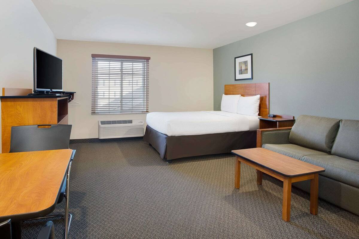 A hotel room with a bed, a desk, a chair, a television, a window with blinds, a sofa, and a picture on the wall.