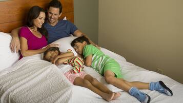 A family of three on a bed with a wooden headboard, a white textured bedspread, and a plain wall in the background.