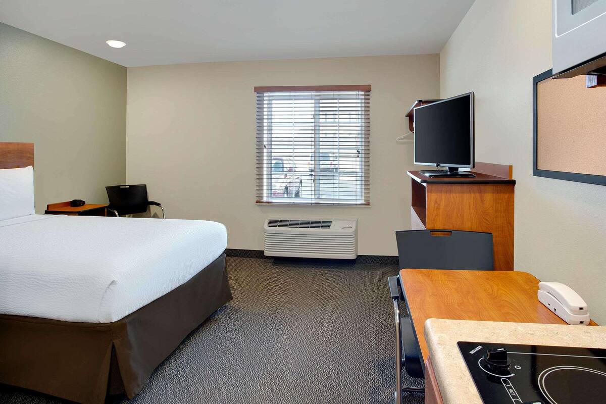 A hotel room with a bed, a desk with a computer, a television, and a window with blinds.