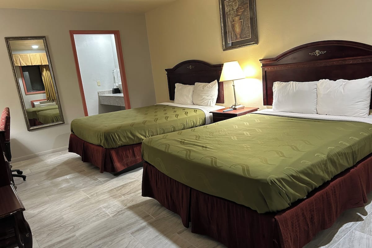 A hotel room with two single beds, a wooden headboard, a nightstand with a lamp, a mirror, and a framed picture on the wall.