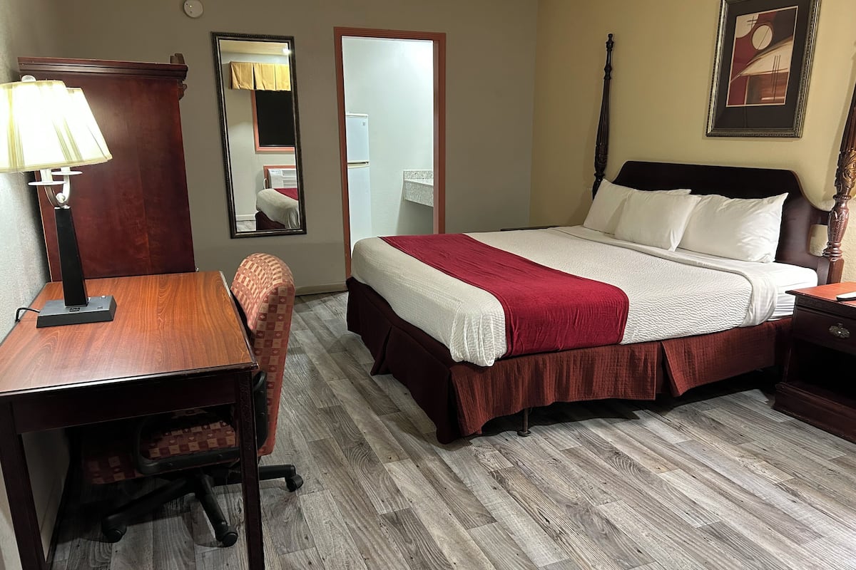 A hotel room with a wooden desk, a chair, a bed with a red and white bedspread, a nightstand, a lamp, a mirror, and a framed picture on the wall.
