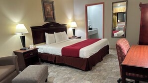 A hotel room with a bed, two bedside tables with lamps, a chair, a small table, and a mirror. - Howards Inn Motel (Eunice)