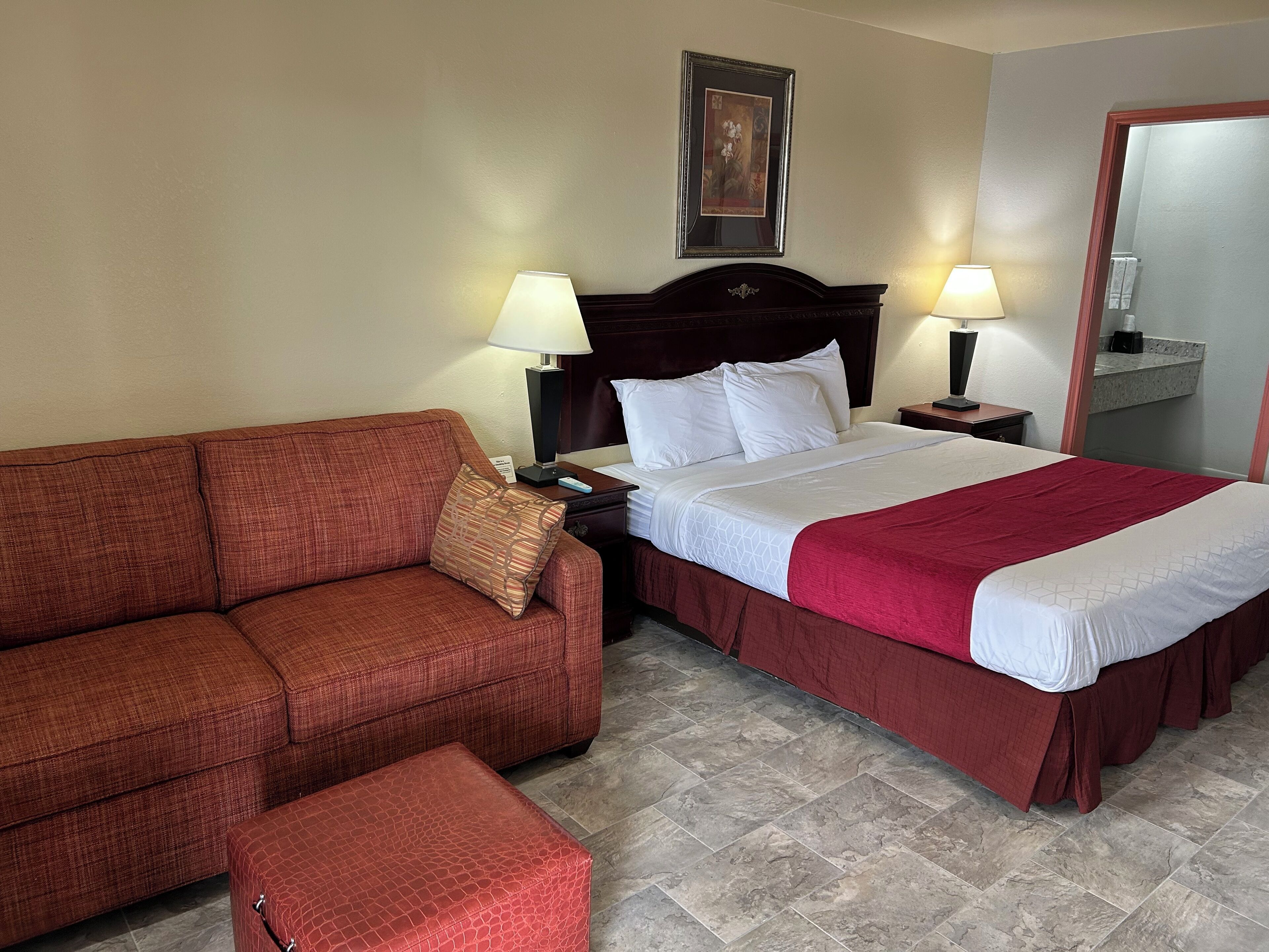 A hotel room with a red sofa, a bed with a red and white bedspread, two bedside lamps, a nightstand, and a framed picture on the wall.