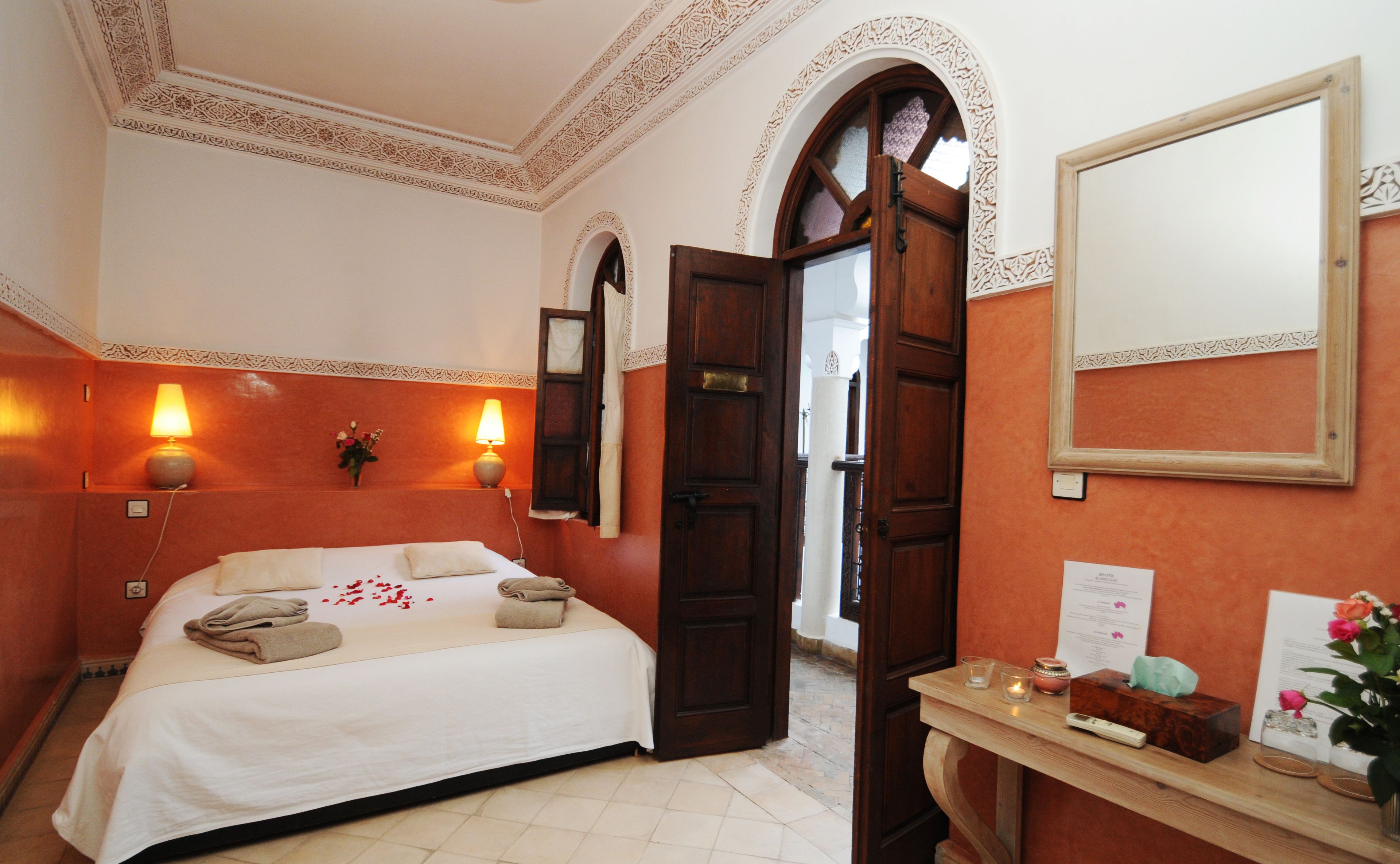Quarto de hotel com cama, duas luminárias fixadas na parede, porta de madeira, espelho, mesa pequena com um vaso de flores e chão em mosaico.