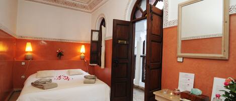 Quarto de hotel com cama, duas luminárias fixadas na parede, porta de madeira, espelho, mesa pequena com um vaso de flores e chão em mosaico.