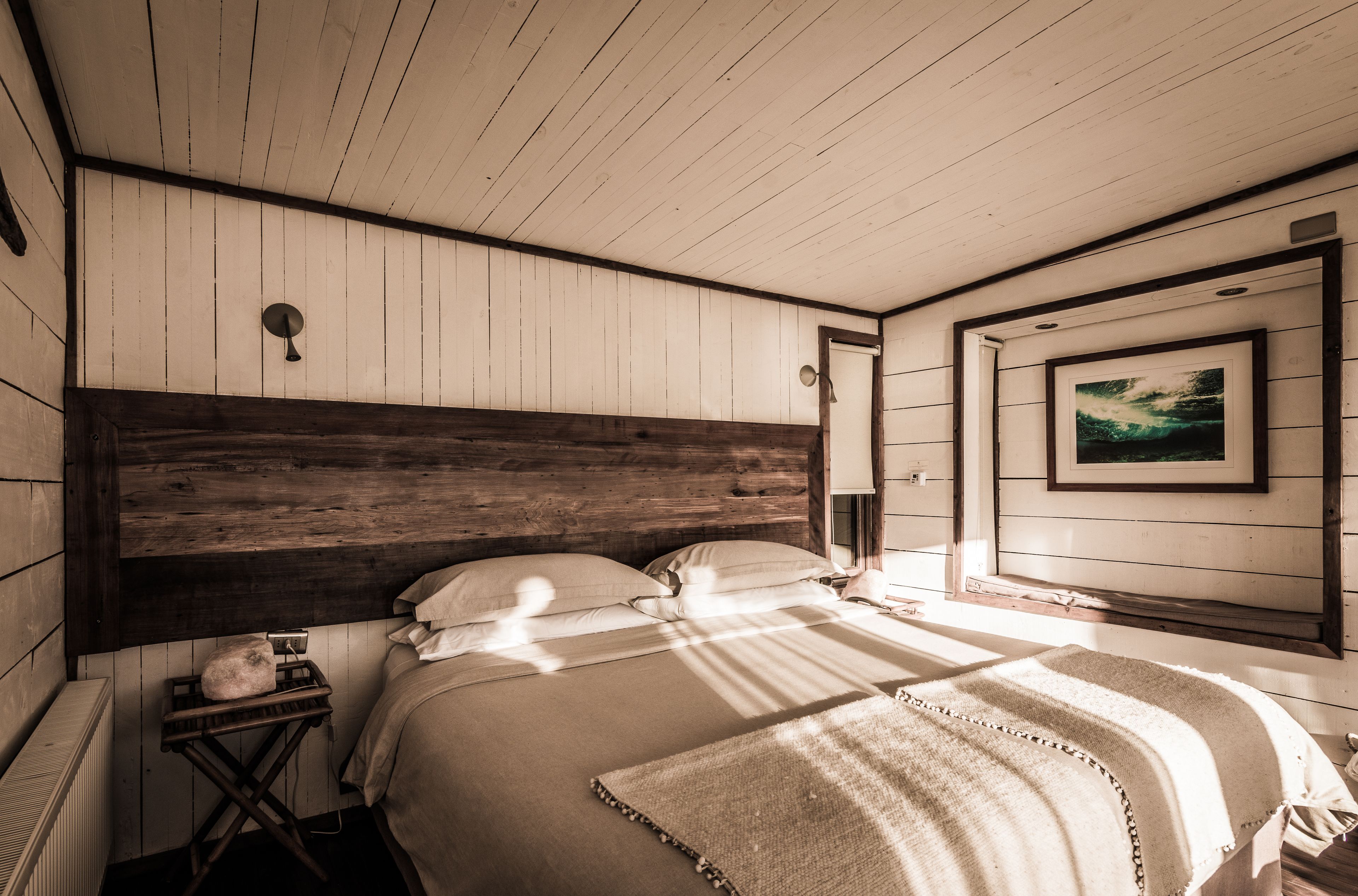 A bedroom with a wooden headboard, a bed with white bedding, a small bedside table with a lamp, and a framed picture on the wall.