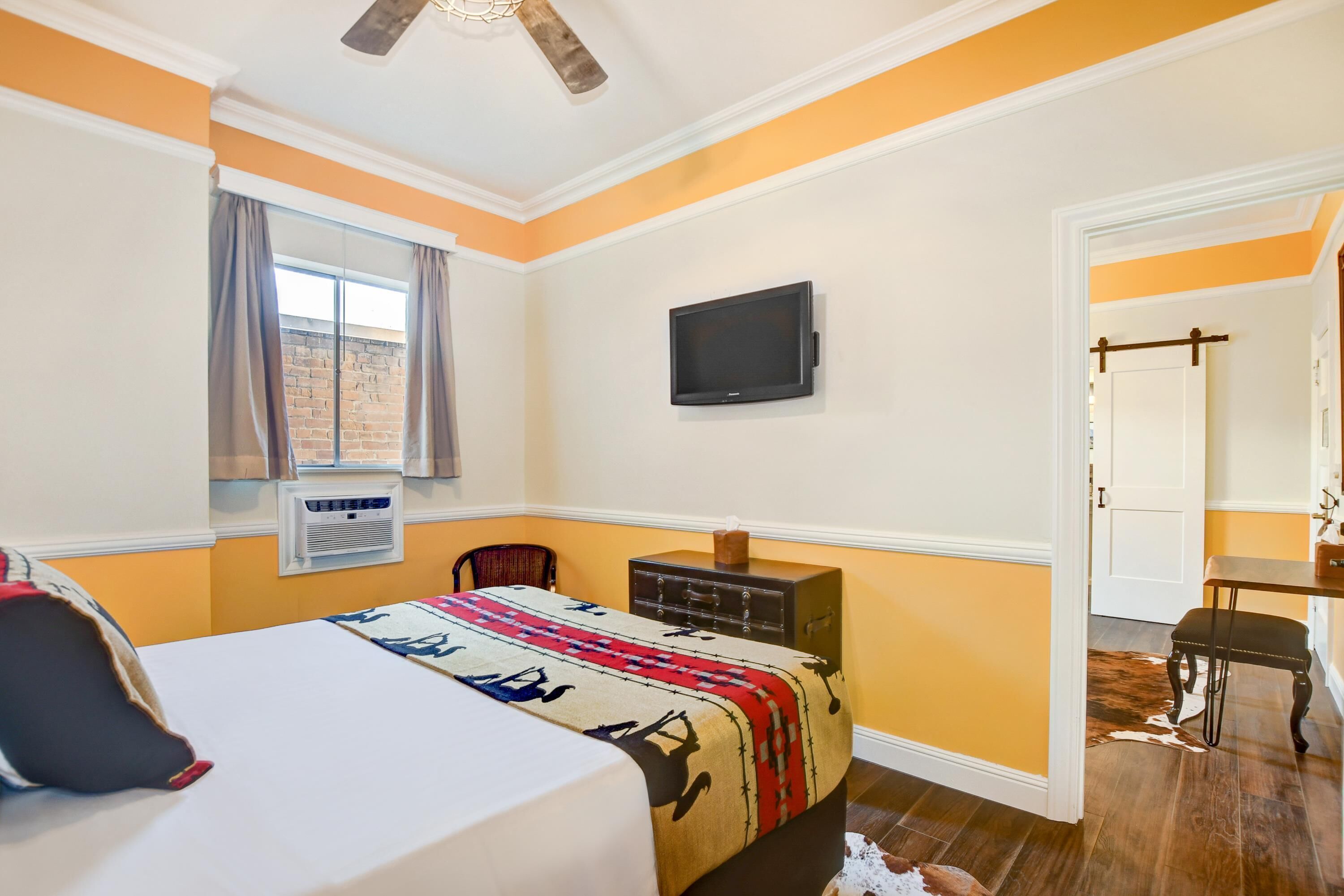 Habitación de hotel con cama, televisión, cómoda, ventana con cortinas y puerta que conduce a otra habitación.