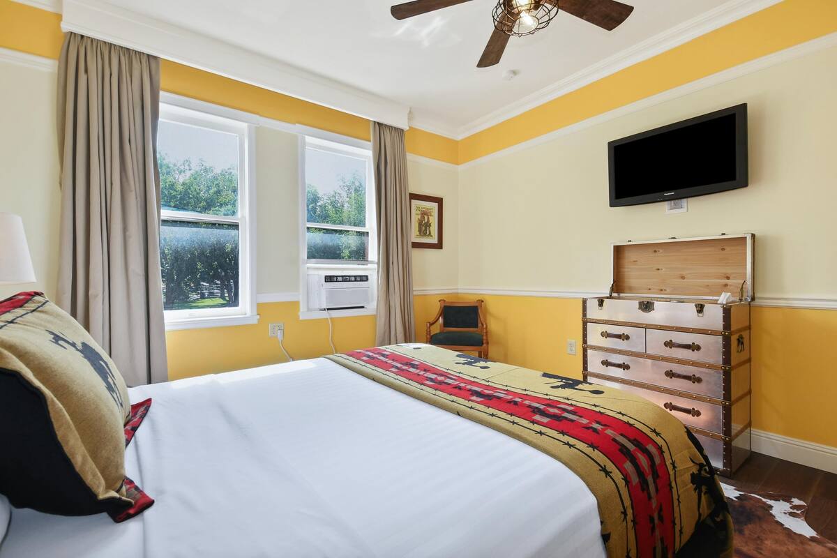 Un dormitorio con cama, televisor, cómoda y ventana con cortinas.